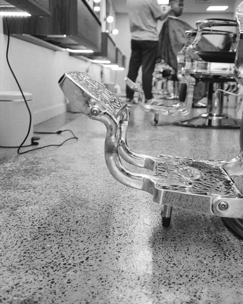 A chrome chair base on the terrazzo floor, barbers at work behind it, in black and white