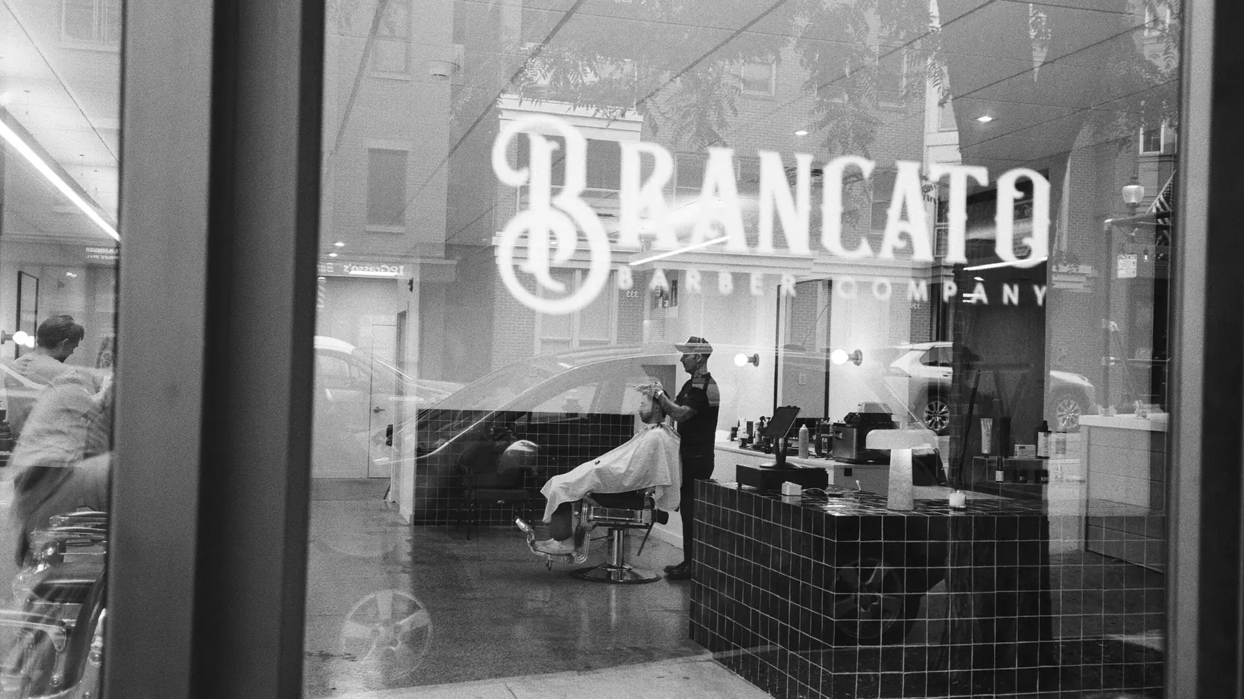 The South Boston shop through its front window after dark, a barber mid-cut inside, in black and white